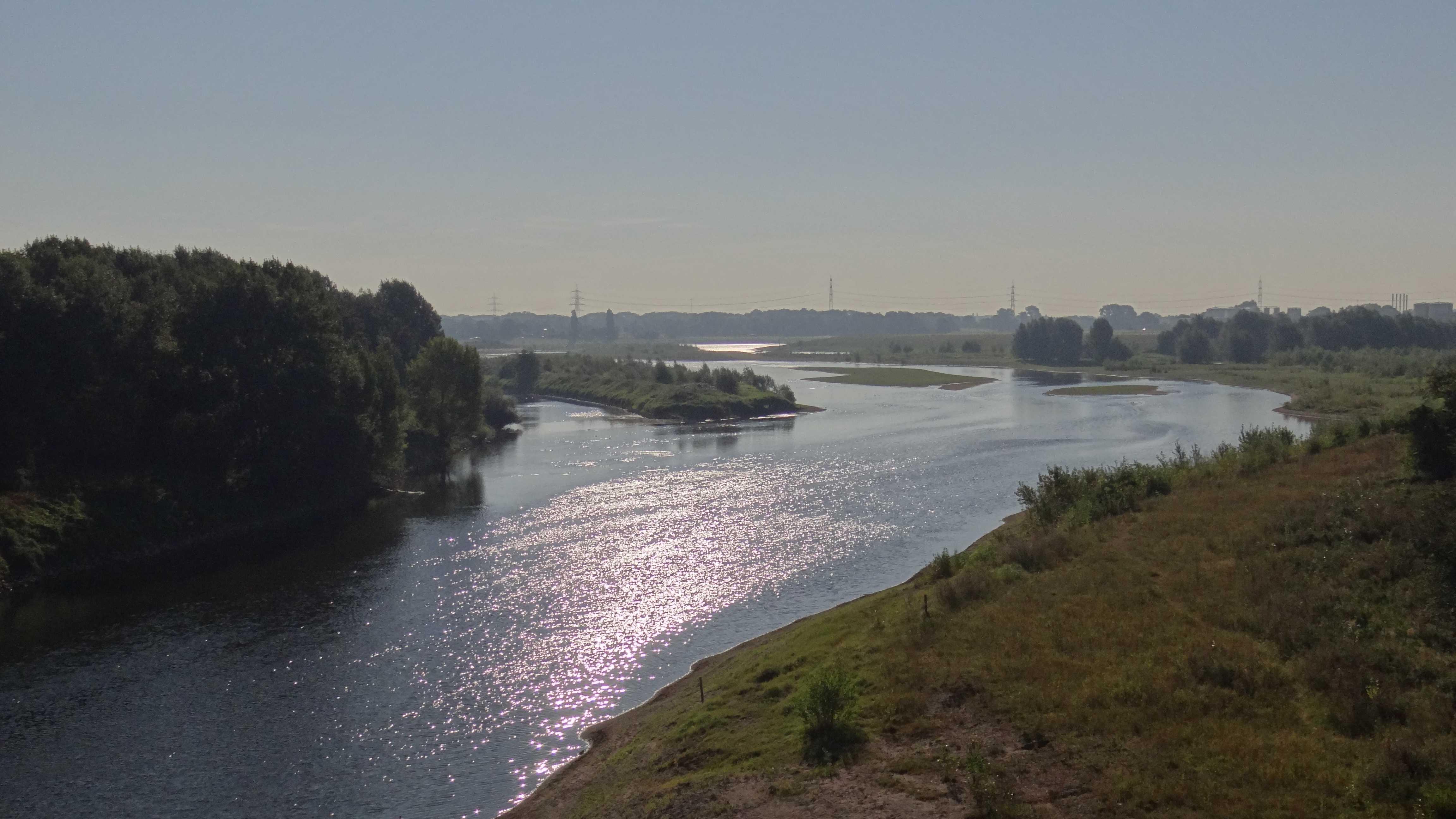 Breiter träger Fluss in ebenem Gelände, Wiesen, Bäume, Industriegebäude am Horizont, Himmel