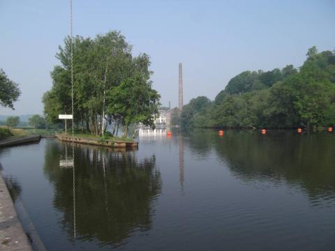 Breiter träger Fluss in ebenem Gelände, Bojenkette, Ufermauern, Bäume, Industriegebäude am Horizont, Himmel