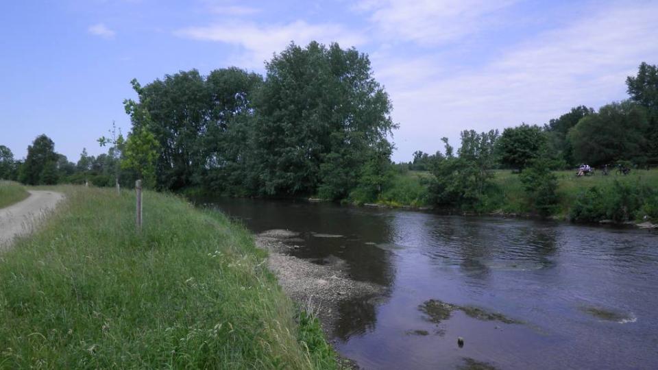 Breiter träger Fluss in ebenem Gelände, Wiesen, Bäume, Himmel