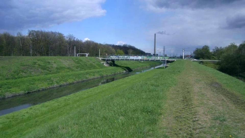 gerader eingedeichter Flusslauf, Gras, Brücke, Baume, Industrieschornstein