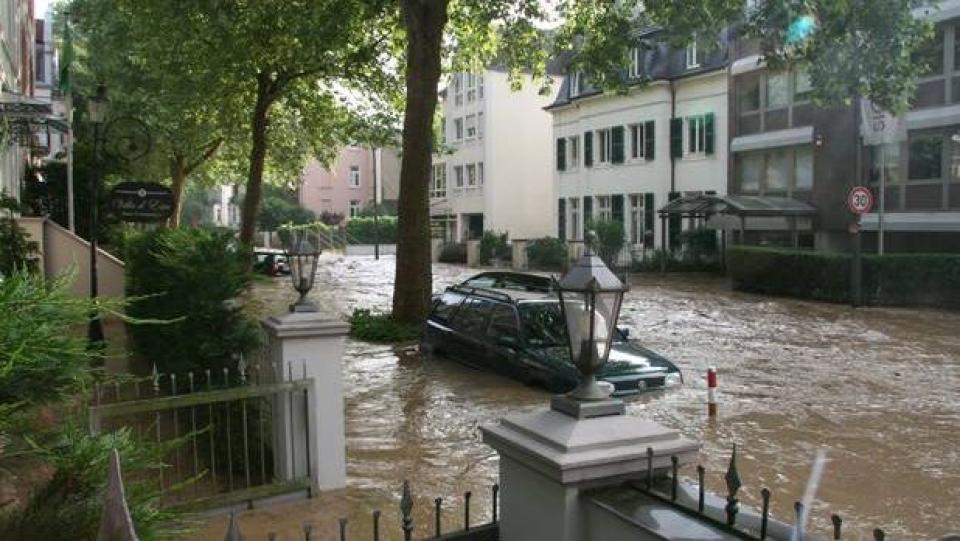 überflutete Straße und Auto, braunes ca. 50 cm tiefes Wasser, aus dem Mauern, Zäune, Bäume, mehrstöckige Häuser ragen, 