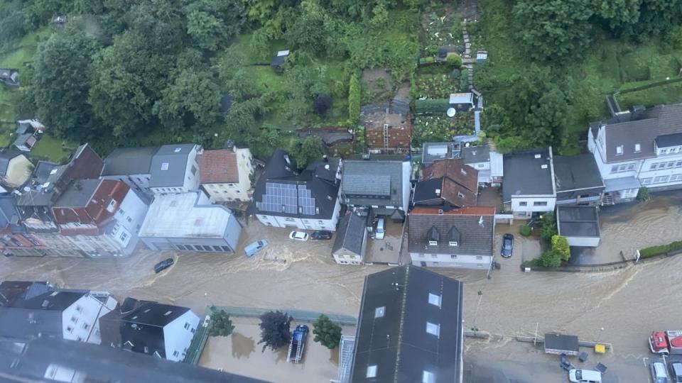 Luftbild einer überfluteten Straße, ca. 50 cm tiefes Wasser, mehrstöckige Häuser, Autos, bewaldeter Hang 