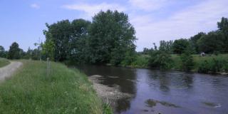 Breiter träger Fluss in ebenem Gelände, Wiesen, Bäume, Himmel