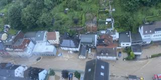 Luftbild einer überfluteten Straße, ca. 50 cm tiefes Wasser, mehrstöckige Häuser, Autos, bewaldeter Hang 