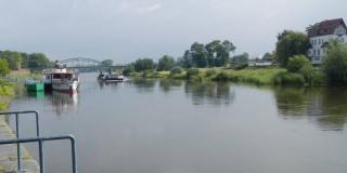 Ein Foto zeigt die Weser, auf der ein Schiff fährt, 2 ankern am linken Ufer. Im Hintergrund ist eine größere Brücke zu sehen, Die Uferböschungen sind teilweise mit Büschen bewachsen, rechts an Land steht ein Haus.