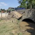 Steinbogenbrücke, Fluss, der schlammig-braunes Wasser führt, angeschwemmtes Holz und zerstörter Zaun am Ufer, Lastwagen, Müllcontainer